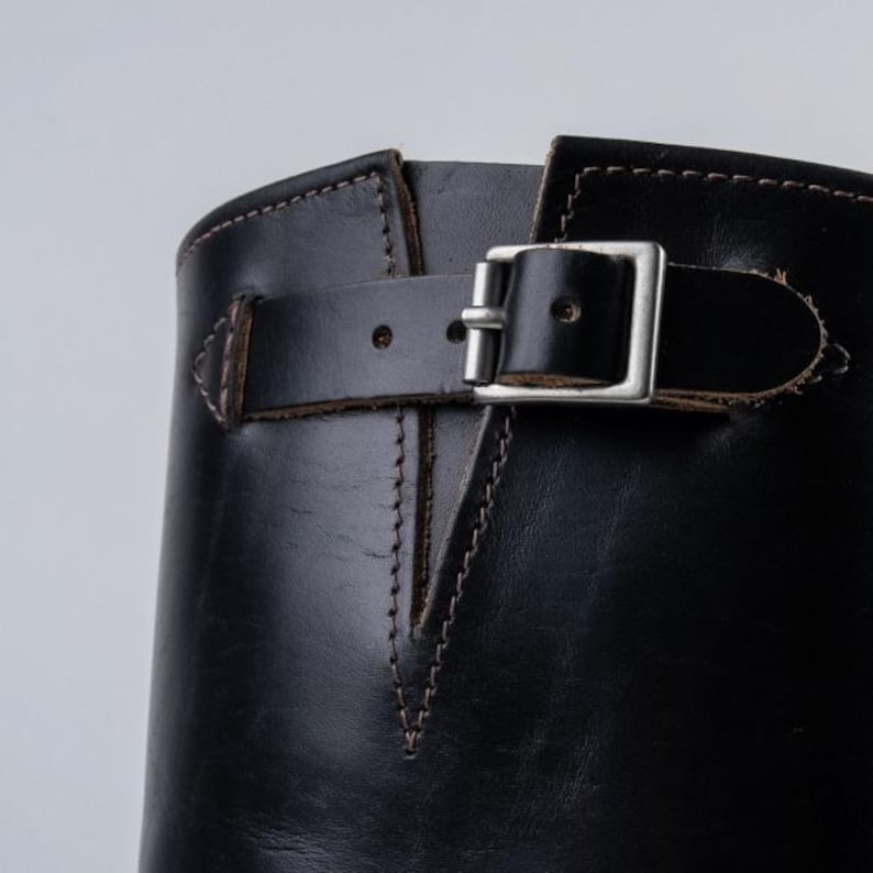 May include: Close-up of a black leather boot with a silver buckle and a brown leather strap. The boot has brown stitching and a smooth, shiny surface. The boot's design is classic and durable.