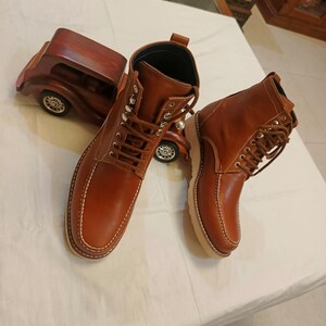 May include: A pair of brown leather boots with white stitching and a lace-up design. The boots have a light-colored sole and are displayed with a wooden car figurine. The boots are a classic style.