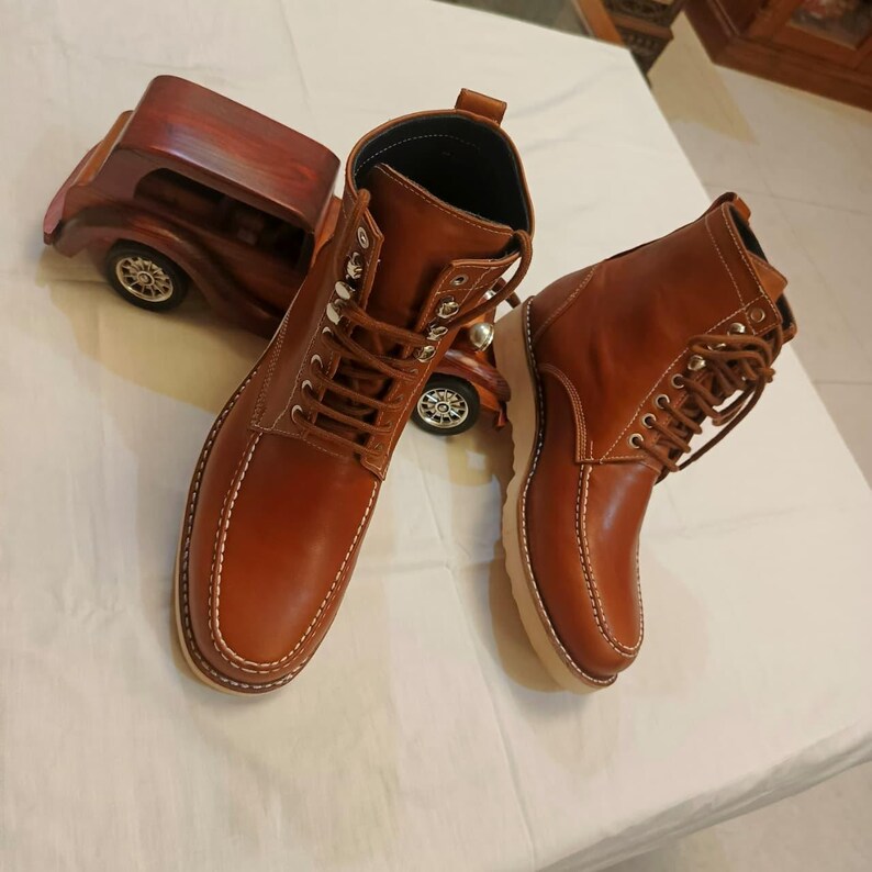 May include: A pair of brown leather boots with white stitching and a lace-up design. The boots have a light-colored sole and are displayed with a wooden car figurine. The boots are a classic style.