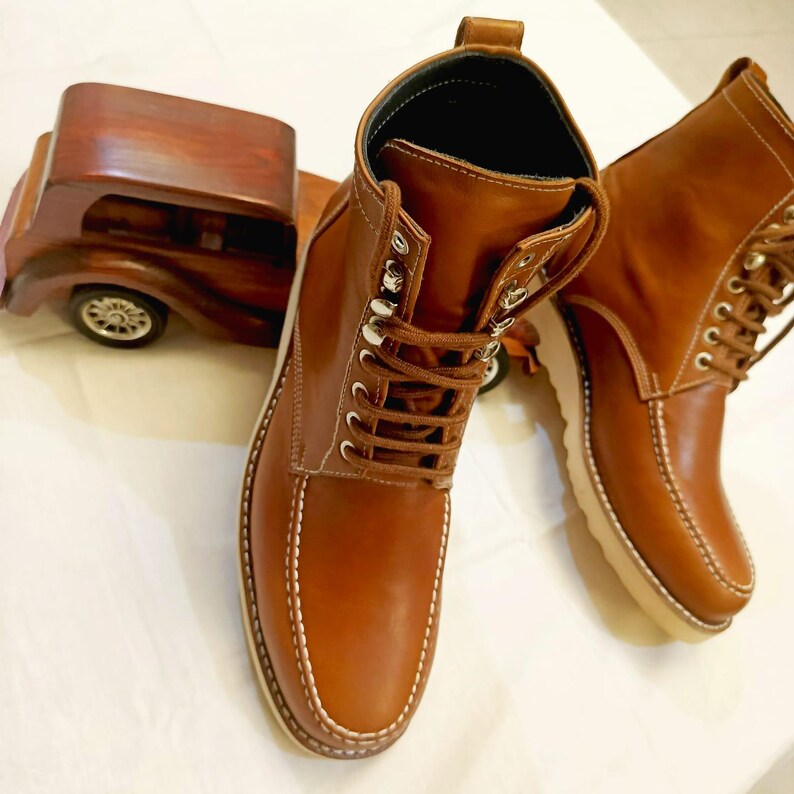 May include: A pair of brown leather boots with white stitching and laces. The boots have a light-colored sole and metal eyelets. A wooden toy car is in the background.