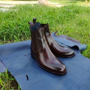 May include: A pair of polished, dark brown leather Chelsea boots. The boots have elastic side panels and pull tabs. They are displayed on a blue surface with green grass in the background.