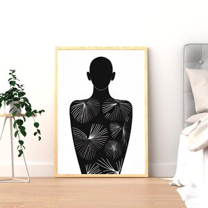 Op de afbeelding: Een ingelijste zwart-wit kunstprint met een silhouet van een persoon. Het lichaam is versierd met witte radiale patronen. Het kunstwerk wordt getoond in een kamer met een plant en een bed.