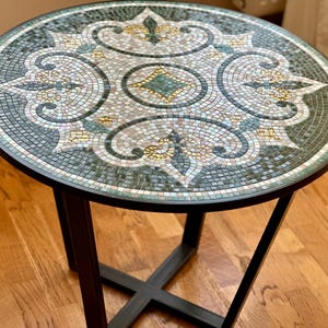 Könnte beinhalten: Runder Beistelltisch mit einer Mosaikfliesenplatte mit einem detaillierten Blumenmuster in Grün-, Weiß- und Goldtönen. Der Tisch hat ein schwarzes Metallgestell mit X-förmigem Fuß, geeignet für Wohn- oder Schlafzimmer.