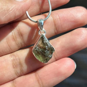 May include: A silver necklace with a raw, olive-green moldavite pendant. The irregularly shaped gemstone is set in a silver bezel and hangs from a delicate silver chain. The pendant's surface has a textured appearance, showcasing its natural form.
