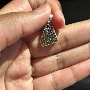 May include: A green and silver pendant. The pendant has a rough, textured green stone with a silver bail. The stone has a unique, organic shape and is held in a silver setting. The pendant is held in a hand.