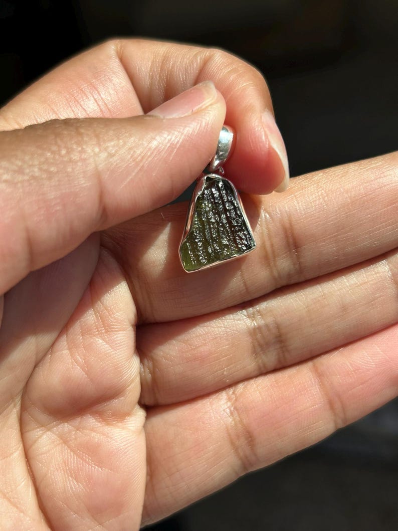 May include: A green and silver pendant. The pendant has a rough, textured green stone with a silver bail. The stone has a unique, organic shape and is held in a silver setting. The pendant is held in a hand.