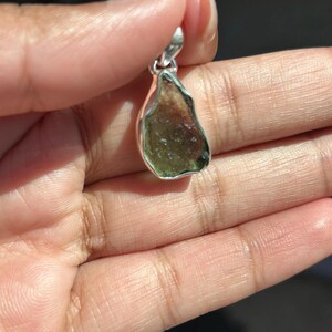 May include: A natural, raw-cut gemstone pendant with a silver-coloured bail. The pendant features a mix of green and brown hues. The stone is held up by a hand.
