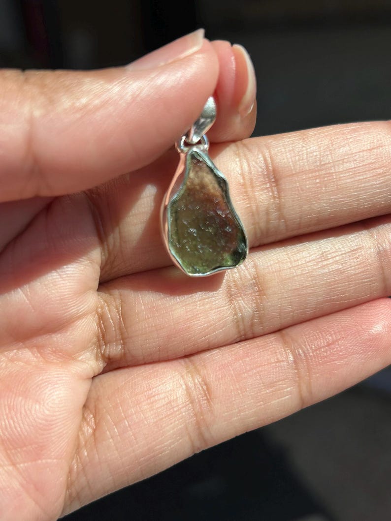 May include: A natural, raw-cut gemstone pendant with a silver-coloured bail. The pendant features a mix of green and brown hues. The stone is held up by a hand.