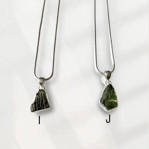 May include: Two silver necklaces with pendants. Each pendant features a rough-cut, dark green stone. The necklaces hang against a white backdrop. The pendants are labelled with the letters "I" and "J".
