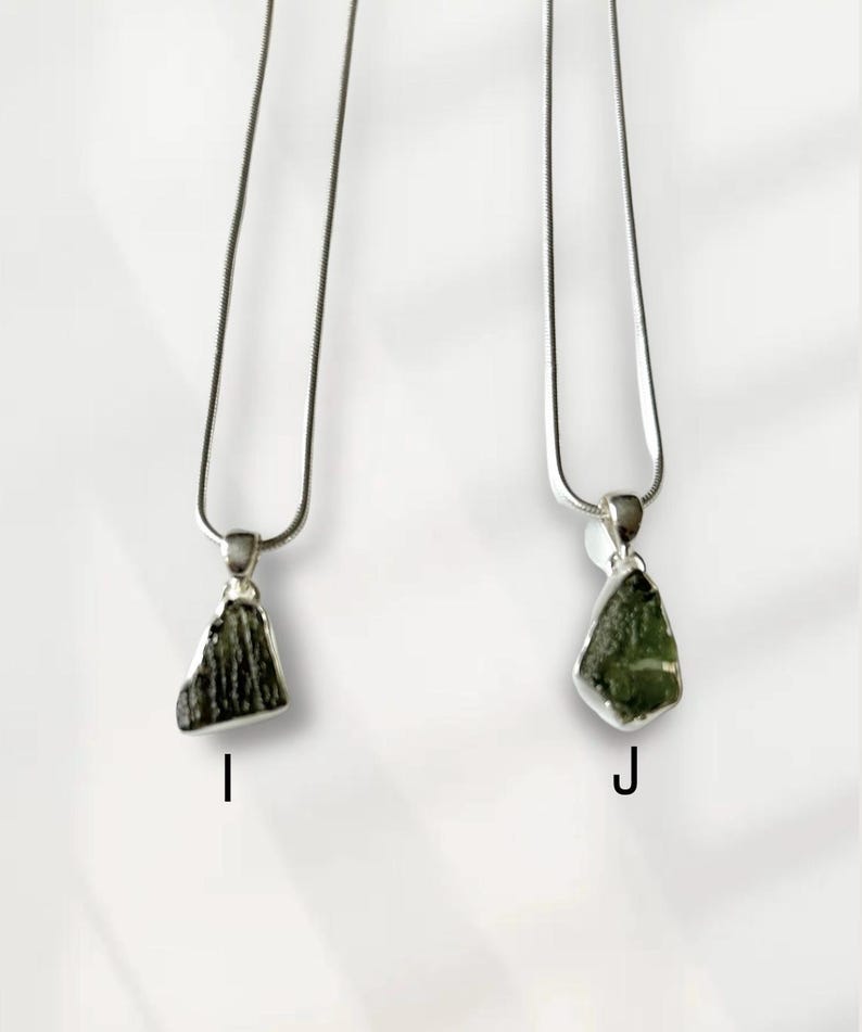 May include: Two silver necklaces with pendants. Each pendant features a rough-cut, dark green stone. The necklaces hang against a white backdrop. The pendants are labelled with the letters "I" and "J".