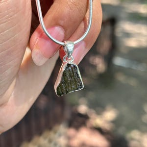 May include: A silver necklace with a pendant made from a rough, dark green gemstone. The pendant is an irregular shape and hangs from a fine silver chain. The necklace is held up by a hand.