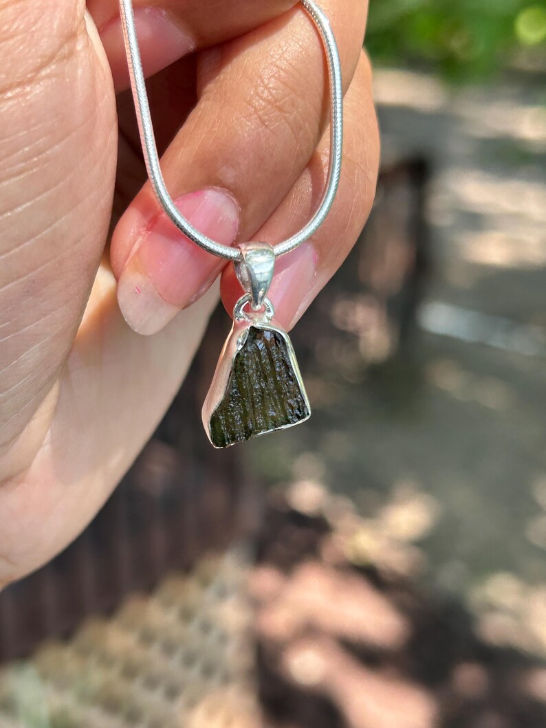 May include: A silver necklace with a pendant made from a rough, dark green gemstone. The pendant is an irregular shape and hangs from a fine silver chain. The necklace is held up by a hand.