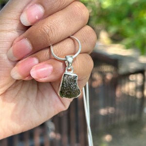 May include: A silver necklace with a pendant showcasing a rough, olive-green gemstone. The pendant has an irregular shape and is set in a silver bezel. The necklace is held against a blurred green backdrop.