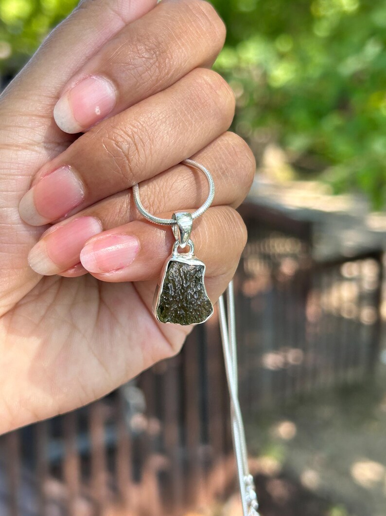 May include: A silver necklace with a pendant showcasing a rough, olive-green gemstone. The pendant has an irregular shape and is set in a silver bezel. The necklace is held against a blurred green backdrop.