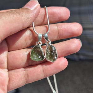 May include: Two pendants with rough-cut, olive-green gemstones set in silver. The pendants are suspended from a delicate silver chain. The gemstones have a natural, textured appearance, and the silver settings have a simple, elegant design.