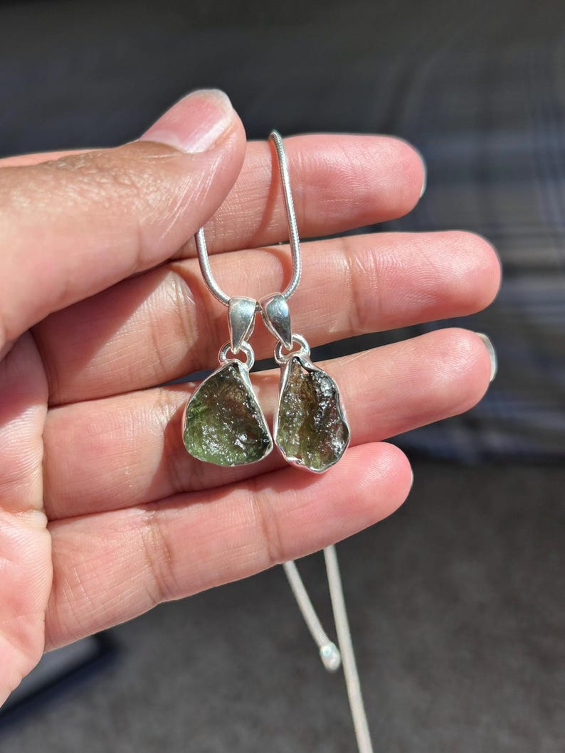 May include: Two pendants with rough-cut, olive-green gemstones set in silver. The pendants are suspended from a delicate silver chain. The gemstones have a natural, textured appearance, and the silver settings have a simple, elegant design.