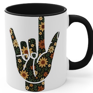 May include: White ceramic mug with a black interior and handle. The mug features a floral design in the shape of the ASL sign for "I love you." The floral pattern includes yellow, orange, and brown flowers on a dark green background.