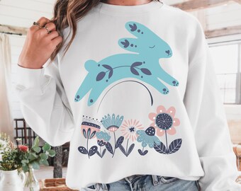 May include: White sweatshirt featuring a light blue bunny leaping over a floral design. The bunny has dark purple accents and the flowers are in shades of blue, pink, and purple. The sweatshirt is a casual, comfortable style.