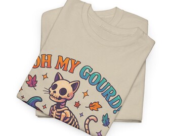 May include: Two beige t-shirts, folded and stacked. The top shirt displays a colourful graphic of a skeleton cat with the text "OH MY GOURD!" surrounded by autumn leaves and stars. The design has orange, blue, and purple accents.