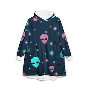 May include: A dark blue hooded blanket featuring a pattern of pink, teal, and blue alien faces, UFOs, and stars. The blanket has a white fleece lining and cuffs, and a large front pocket. The design is repeated across the entire blanket.