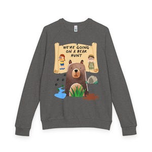 Peut inclure: Un sweat-shirt gris anthracite à manches longues avec une impression graphique représentant un ours brun, deux enfants avec des jumelles, une grotte, une rivière et des empreintes de pattes. Le texte "WE'RE GOING ON A BEAR HUNT" est inscrit sur un parchemin beige.