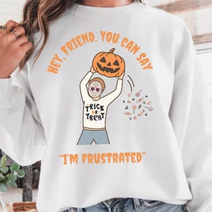May include: White Halloween sweatshirt with a cartoon illustration of a person wearing a mask and holding a jack-o'-lantern. The sweatshirt has the text "Trick or Treat" and "I'm Frustrated". The words "Hey, Friend, You Can Say" are printed above the pumpkin.