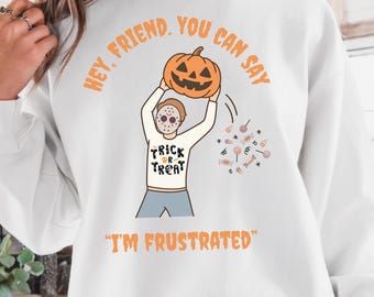 May include: White Halloween sweatshirt with a cartoon illustration of a person wearing a mask and holding a jack-o'-lantern. The sweatshirt has the text "Trick or Treat" and "I'm Frustrated". The words "Hey, Friend, You Can Say" are printed above the pumpkin.