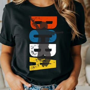 May include: Black t-shirt featuring the word "BOBA" in large, distressed letters. The letters are in orange, blue, white, and yellow. The shirt is a casual style.