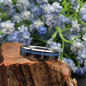 May include: A silver-colored ring with a thin, bright blue stripe. The ring is set on a rough, reddish-brown stone, with a backdrop of small, blue and white flowers.