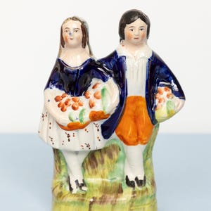 May include: A ceramic figurine depicting a couple in traditional attire. The woman wears a blue shawl and a white dress with floral accents, holding a basket of fruit. The man is in a blue jacket and orange pants.