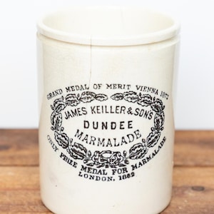 May include: A cylindrical off-white ceramic jar featuring black printed text that reads: GRAND MEDAL OF MERIT VIENNA 1873, JAMES KEILLER & SON'S DUNDEE MARMALADE, ONLY PRIZE MEDAL FOR MARMALADE LONDON, 1862. The jar has a visible vertical crack on the front.