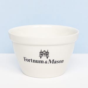 May include: A cream-colored ceramic mixing bowl with a wide, open top and a tapered base. The front of the bowl features a black printed logo with an ornate clock tower illustration above the text Fortnum & Mason in a classic serif font.