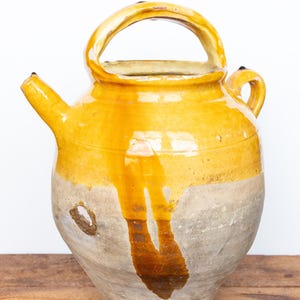May include: A bulbous ceramic pitcher with a mustard yellow glazed upper section and a raw, unglazed grey lower body. The vessel features a prominent arched top handle, a smaller side handle, and a short, angled spout with visible drip marks.