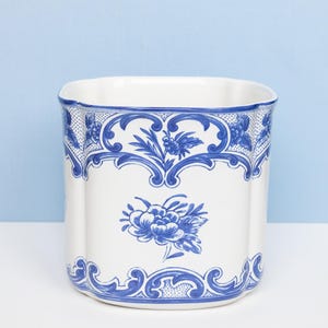 May include: A white ceramic planter with a scalloped rim and a blue floral pattern. The design features intricate scrollwork borders at the top and bottom, with a central blue flower motif on the front. The vessel has a rounded square shape.