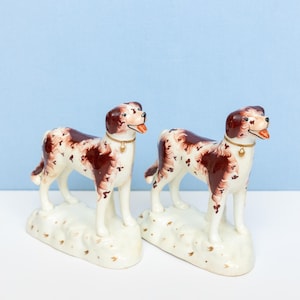 May include: A pair of ceramic dog figurines featuring a white base with brown patches and spots. Each dog stands on a textured white platform, wears a gold-colored collar with a small bell, and has an open mouth showing a red tongue.