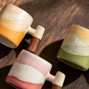Könnte beinhalten: Drei Keramikbecher mit Holzgriffen. Zwei Becher haben einen gelb-orangen oder grünen Farbverlauf mit dem eingravierten Wort DAD und einem Herz. Ein Becher hat einen rosa-weißen Farbverlauf mit dem eingravierten Wort MOM und einem Herz.