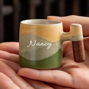 Può includere: Una piccola tazza in ceramica con un design a strati colorati nelle tonalità del giallo, verde chiaro e verde scuro. La tazza ha un corpo cilindrico con il nome Nancy stampato in bianco e un manico con un'estensione in legno marrone scuro.