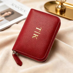 Puede incluir: Una cartera de cuero rojo intenso con textura granulada, cierre de cremallera dorado y tirador de cuero a juego. La parte frontal presenta las letras TIK doradas en relieve, centradas verticalmente en una tipografía con remates.