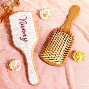 May include: Two wooden hairbrushes on a pink surface. One brush has a white handle with the name "Marry" in pink rhinestones. The other brush is made of wood with brown bristles.
