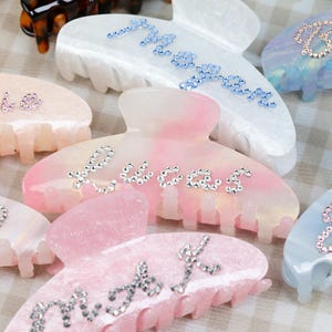 May include: A selection of hair claw clips in pastel shades. Each clip features rhinestone lettering, spelling out names such as "Lucas", "Moon", and "Bee". The clips are available in pink, blue, and white, offering a range of options.
