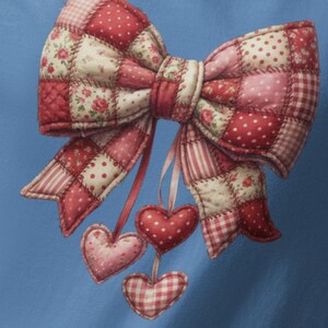 Könnte beinhalten: Hellblaues Langarmshirt mit einem großen Patchwork-Schleifen-Design. Die Schleife ist hauptsächlich rot und weiß, mit Blumen- und Vichy-Mustern. Drei herzförmige Ornamente hängen darunter und verleihen einen Hauch von Laune.