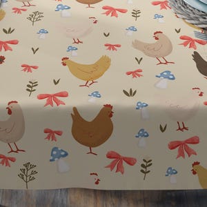 Peut inclure: Une nappe beige avec un motif répété de poulets, de nœuds rouges, de champignons bleus et de brins verts. La nappe est posée sur une table en bois, avec des couverts et des verres à vin visibles.