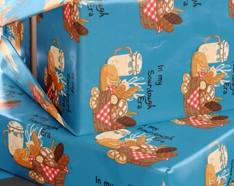 Puede incluir: Cajas de regalo azules envueltas en papel estampado con una ilustración de dibujos animados de pan, una cesta y una jarra. El papel tiene el texto "In my Sourdough Era". También se ven tijeras y cinta.