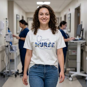 Può includere: Una maglietta bianca con la scritta "NURSE" in lettere a blocchi blu e gialle, con un disegno di stetoscopio. Il testo "Lorianna NICU" è sopra la parola "NURSE". La persona indossa jeans blu.