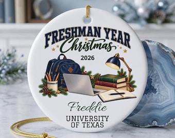 May include: A round white ceramic ornament featuring the text FRESHMAN YEAR Christmas 2026, Freddie, and UNIVERSITY OF TEXAS. The graphic shows a blue backpack, a laptop, a desk lamp, stacked books, and an open notebook with a pen.