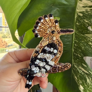May include: A beaded bird brooch featuring a detailed design with gold, black, white, and iridescent beads. The bird is perched on a brown branch, with a fan-shaped crest. The brooch is likely handmade and suitable for adding to clothing or accessories.