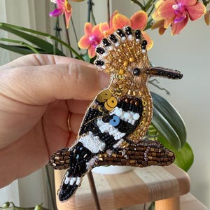 May include: A handcrafted bird brooch with intricate beadwork. The brooch features a detailed design with gold, black, white, and blue beads, creating a realistic depiction of a bird. The bird is perched on a brown beaded branch.