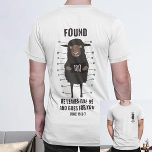Puede incluir: Camiseta blanca con un diseño de oveja negra y la palabra "FOUND" encima. La oveja está colocada contra un fondo de alineación policial, sosteniendo un letrero que dice "100". El texto "HE LEAVES THE 99 AND GOES FOR YOU LUKE 15:3-7" está debajo.