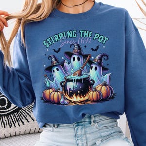 Könnte beinhalten: Ein blaues Sweatshirt mit einem Halloween-Motiv. Das Design zeigt drei Geister mit Hexenhüten, die einen Kessel umrühren. Der Text "Stirring the Pot Since 1692" befindet sich über den Geistern, mit Kürbissen um den Kessel.