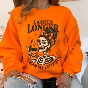 May include: Orange sweatshirt with the text "Lashes Longer Than My Patience" and a skeleton graphic holding a coffee cup. The skeleton has a bow and a headband. The sweatshirt is a solid orange color.